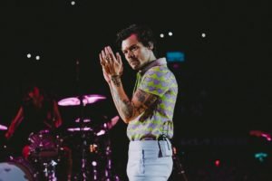 O cantor Harry Styles, um homem jovem, branco, com cabelo liso castanho posa para foto em um palco durante um show. Ele usa uma camisa polo lilás e verde e uma calça de couro também lilás. - Metrópoles
