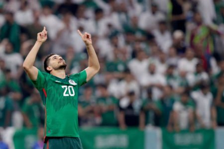 Homem com camisa verde do México com as mãos para cima comemorando um gol marcado na copa do mundo. Ao fundo aparece uma torcida vestida em roupas verdes e brancas