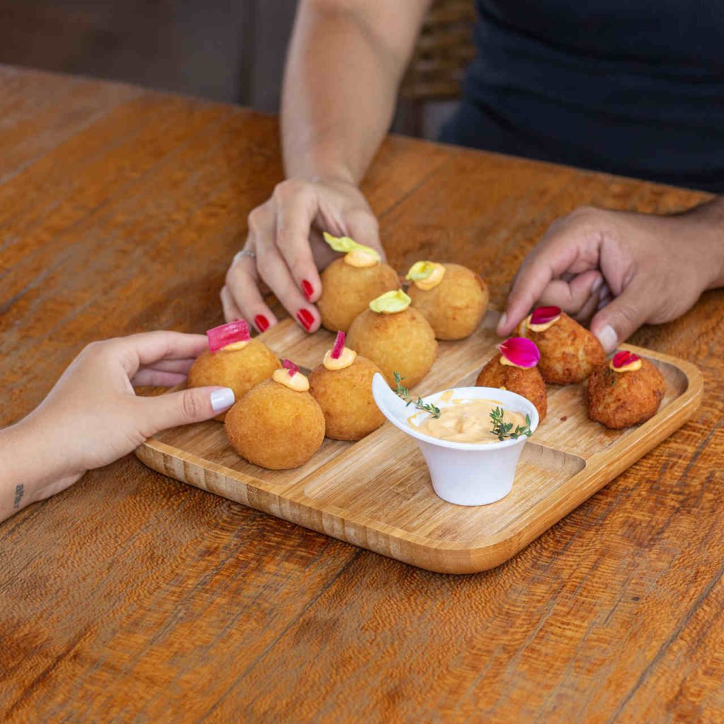 Na foto, duas pessoas dividem uma tábua de petiscos com bolinhos fritos e molhos - Metrópoles