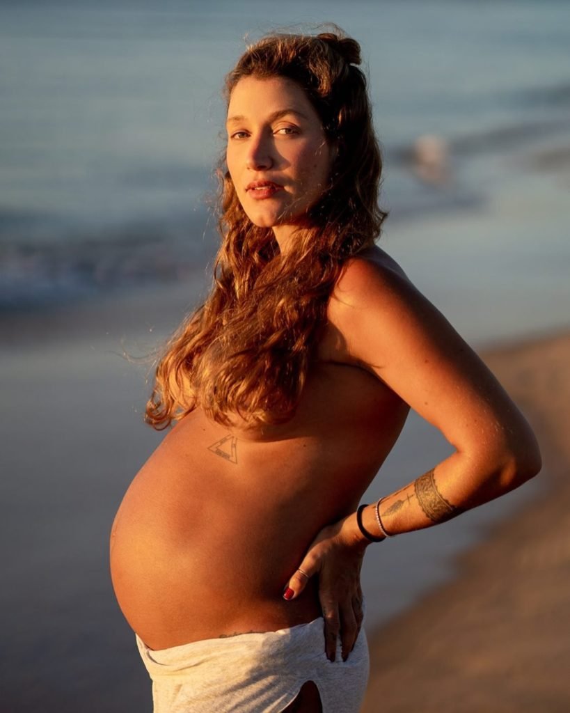 A influenciadora Gabriela Pugliesi, uma mulher branca, jovem e com cabelo ondulado castanho, posando para foto na praia. Ela está grávida e não usa nenhuma blusa na foto. O cabelo longo tampa os seios e evidencia a barriga. - Metrópoles.