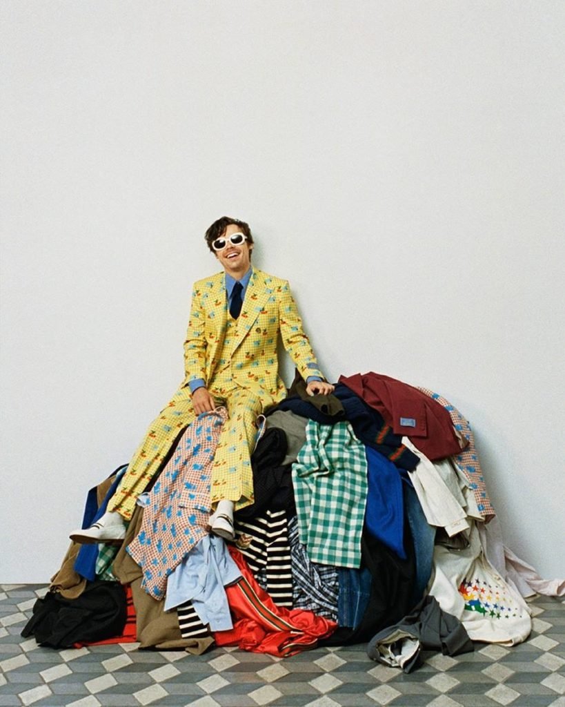 O cantor Harry Styles, um homem jovem e branco, com cabelo curto ondulado, posa para foto em um fundo branco.  Ele usa um terno amarelo e óculos escuros e está sentado em uma pilha de roupa.  A foto faz parte da campanha Gucci Ha Ha Ha, uma coleção da marca italiana com o cantor.  - Metrópoles