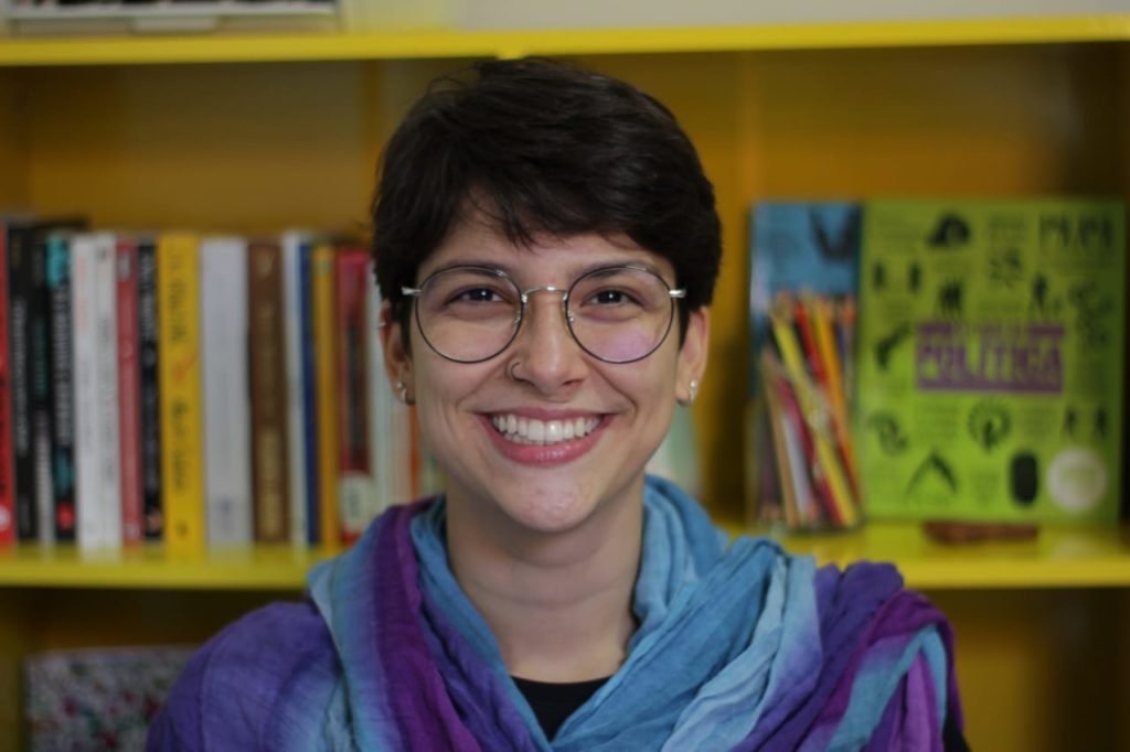 Mulher com cabelo curto e óculos sorrindo em frente a estante de livros