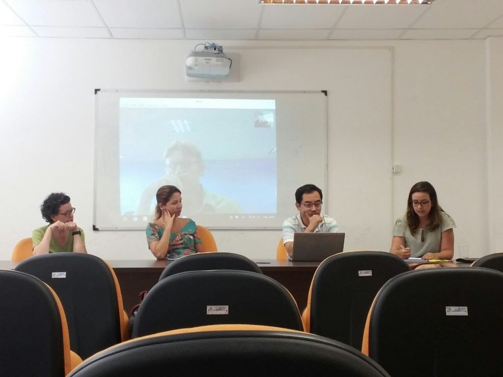 Pessoas sentadas em mesa em frente ao projetor.  Na frente deles há cadeiras vazias