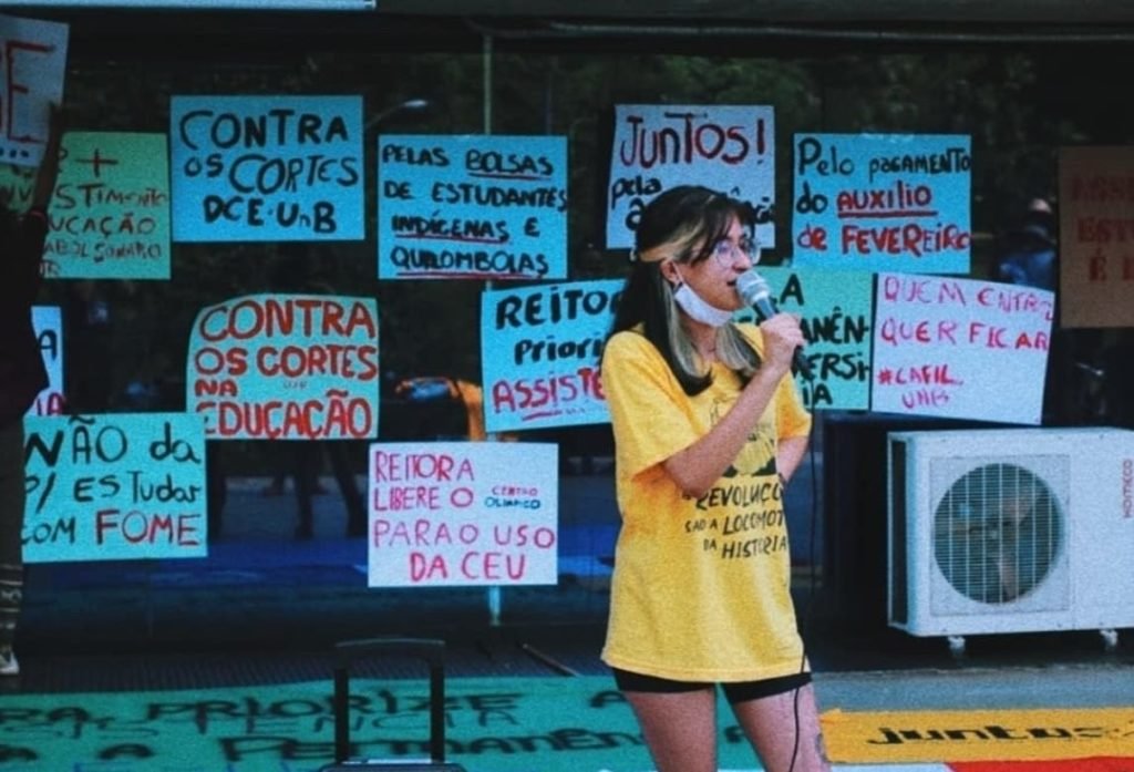 Menina com camisa amarela falando ao microfone em frente a cartazes fixados em vidro escuro