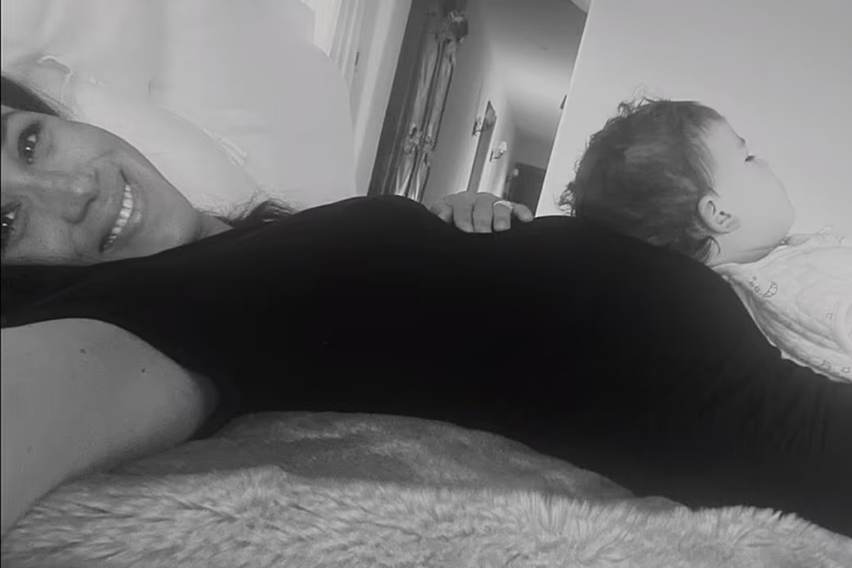 Foto preto e branco de mulher de roupa preta deitada. Ela está grávida e um bebê está apoiado em sua barriga - Metrópoles