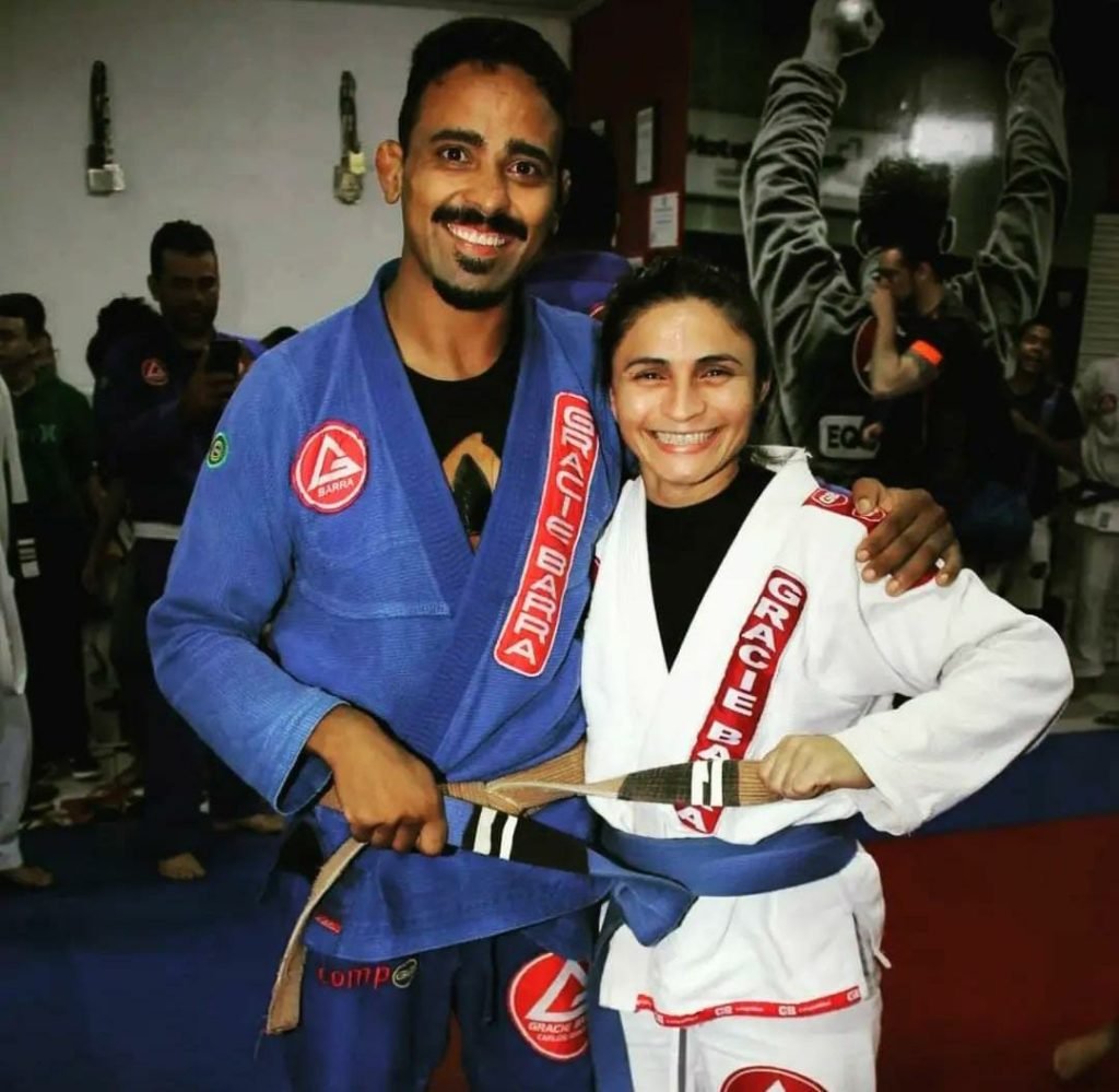 Homem e mulher abraçados, com roupão de luta, em tatame