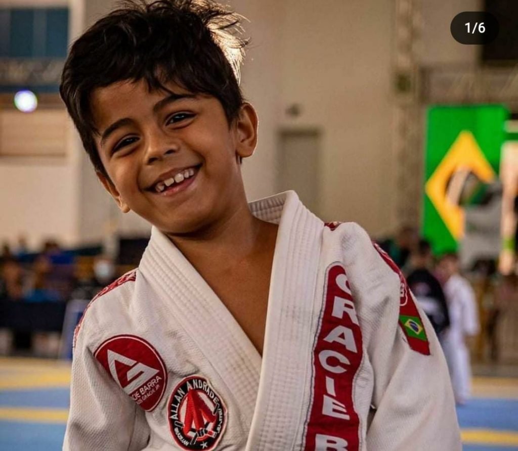 Menino pequeno com roupão branco sorrindo. Há uma bandeira do Brasil ao fundo