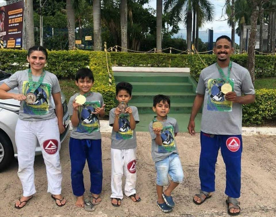 Homem, mulher e crianças ao meio com medalhas em frente a escada. Há árvores ao fundo