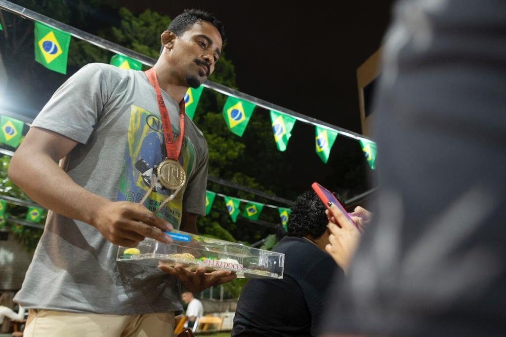 Homem com medalha no pescoço segura bandeja com brigadeiros em bar. Há bandeiras do brasil ao fundo