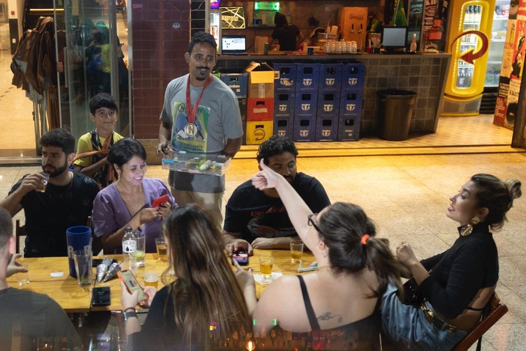 Homem com medalha no pescoço vende brigadeiros para clientes em bar