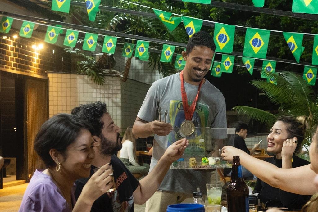 Homem com medalha no pescoço vende brigadeiros para clientes em bar. Há bandeiras do Brasil ao fundo