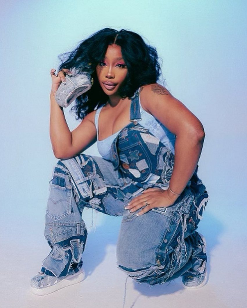 A cantora SZA, uma mulher negra e jovem com cabelo preto ondulado e longo, posa para uma campanha da marca Crocs. Ela usa top azul e um macacão jeans. - Metrópoles