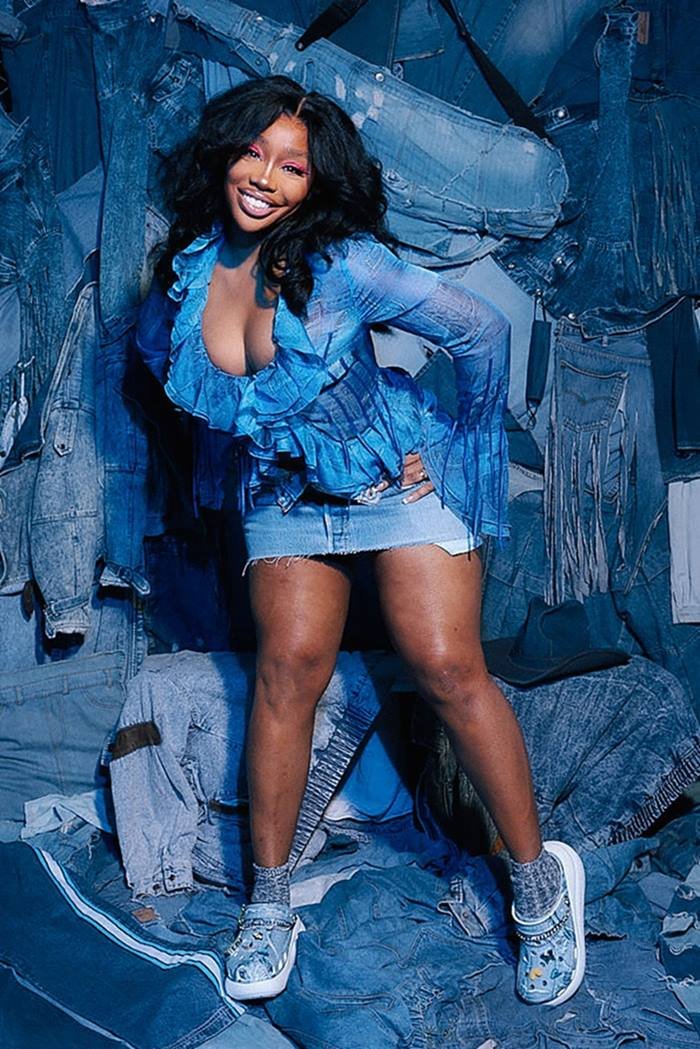 A cantora SZA, uma mulher negra e jovem com cabelo preto ondulado e longo, posa para uma campanha da marca Crocs. Ela usa blusa e calça, ambas em tecido jeans, e o sapato da coleção, uma Crocs, também em jeans. - Metrópoles