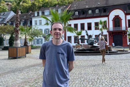 Fábio Almeida em pé, de camiseta azul, na frente de uma casa na Europa - Metrópoles