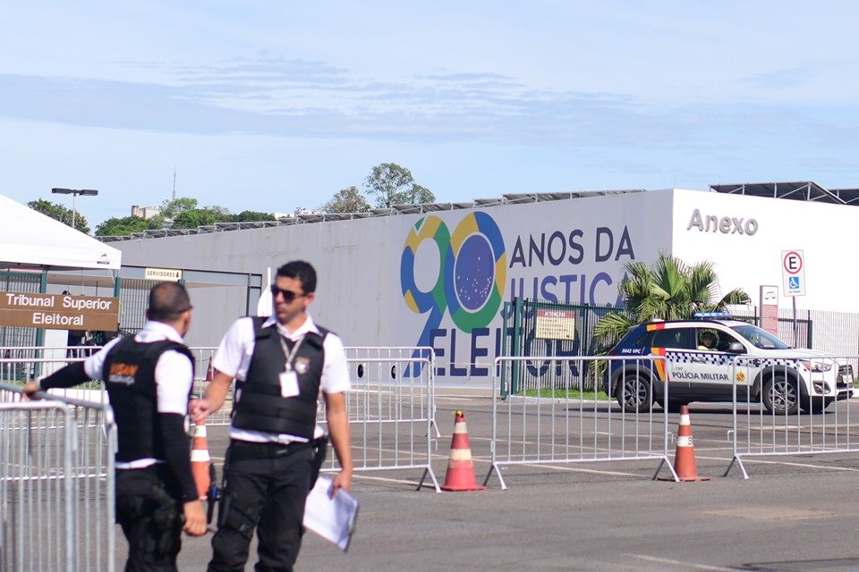 Dois homens uniformizados, de camiseta branca e colete à prova de balas pretos, conversam em frente a placa de identificação do TSE. Ao fundo, um muro branco tem os dizeres