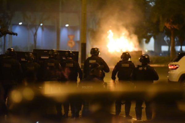 Imagem colorida mostra policiais com escudos, combatendo protesto de bolsonaristas. Carro pega fogo