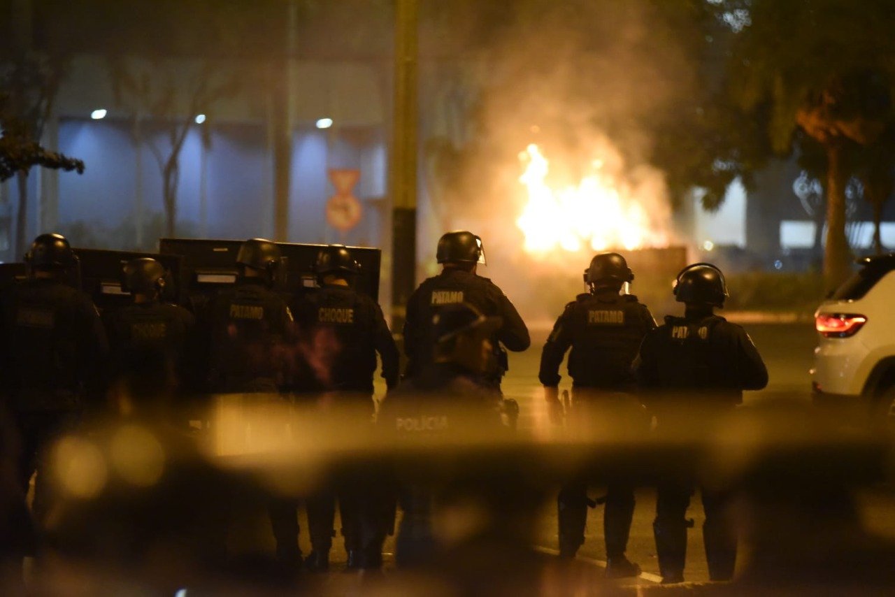 Imagem colorida mostra policiais com escudos, combatendo protesto de bolsonaristas. Carro pega fogo