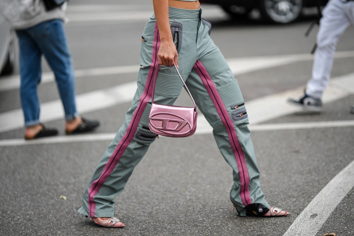 Mulher branca e jovem atravessando a rua. Ela usa uma calça cinza folgada com listras rosa ao lado e segura uma bolsa rosa cintilante da marca Diesel 1DR. - Metrópoles