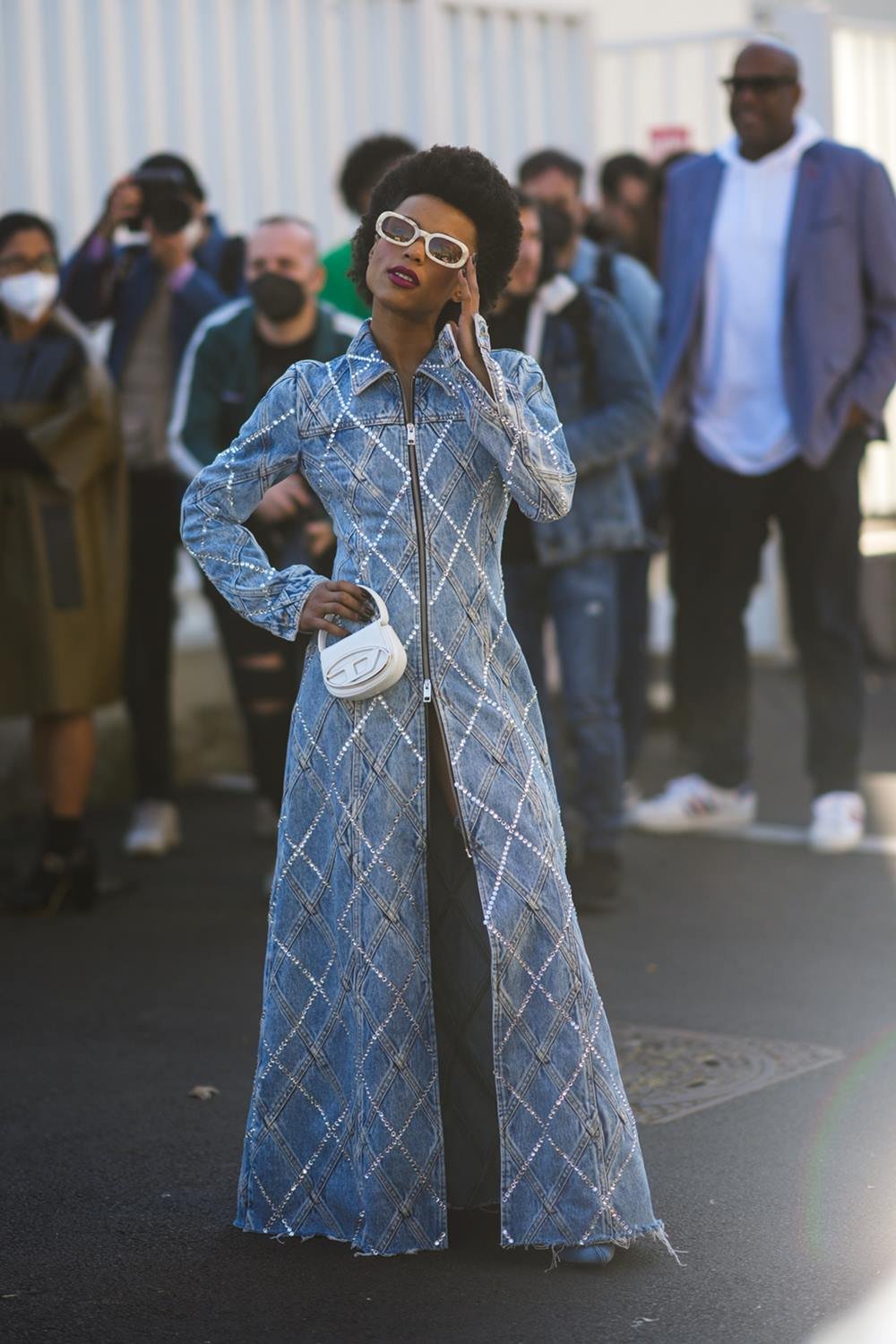 Uma mulher negra e jovem, de cabelos crespos curtos, posa para foto durante a Semana de Moda de Milão. Ela usa um vestido longo jeans, um óculos de armação branca e uma bolsa tamanho mini branca da marca Diesel. - Metrópoles