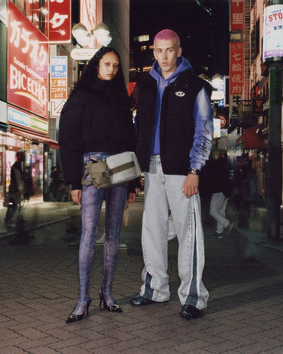 Dois modelos jovens posam para foto em uma rua lotada em Tóquio. A imagem é de uma campanha de divulgação da marca Diesel. A menina é negra, possui o cabelo cacheado longo e usa um moletom preto e uma legging roxa. O homem é branco, tem a cabeça raspada e usa um casaco moletom roxo, um colete preto por cima e calças beges. - Metrópoles