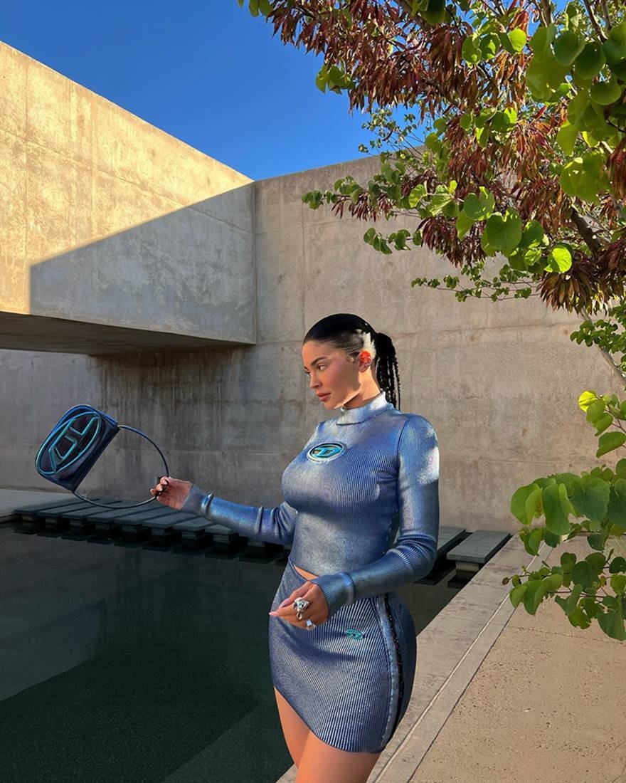 Kylie Jenner, uma mulher branca e jovem, de cabelo longo preto, posa para foto em frente à piscina de sua casa. Ela usa um conjunto de top e saia, ambos azuis cintilantes, e segura uma bolsa modelo 1DR da marca Diesel. A bolsa também é azul cromado. - Metrópoles