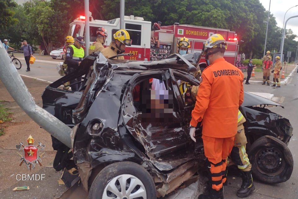 Bombeiro, com uniforme laranja, parado ao lado de ferragens de carro preto danificado após acidente. Ao fundo, caminhão do CBMDF