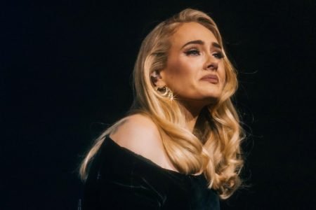 A cantora Adele, uma mulher jovem, branca e com cabelo loiro liso, chorando durante um show. Ela usa um vestido preto e brincos dourados. - Metrópoles