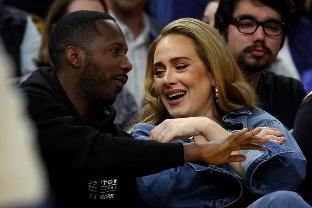 A cantora Adele, uma mulher jovem, branca, com cabelo liso loiro, assiste a um jogo de basquete ao lado de Rich Paul, seu novo namorado. Ele é um homem jovem negro de cabelo raspado curto. - Metrópoles