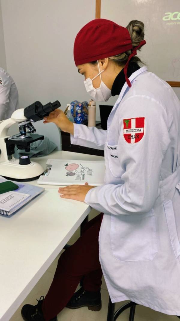 Foto colorida de aluna de Medicina usando touca vermelha e jaleco branco olhando em microscópio-Metrópoles