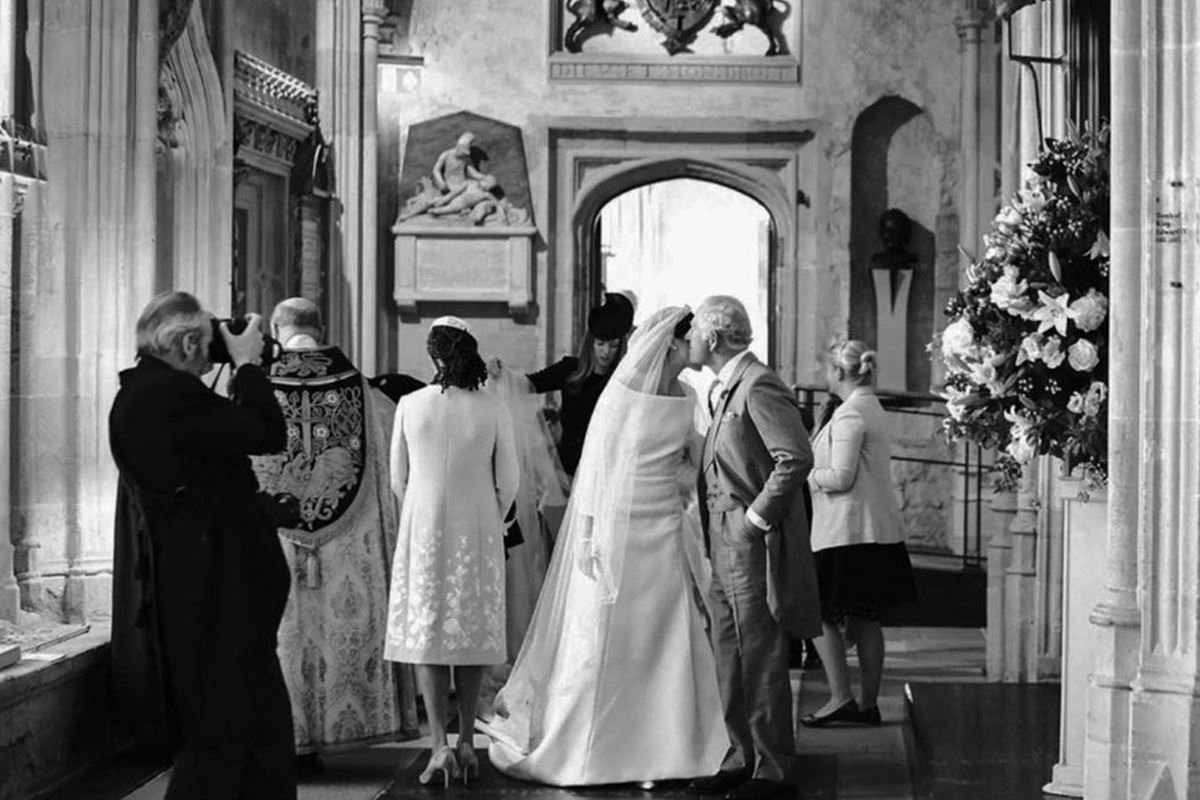 Foto em preto e branco em que mulher vestida de noiva recebe beijo na bochecha de homem vestido de terno. Ao redor deles, pessoas de costas - Metrópoles