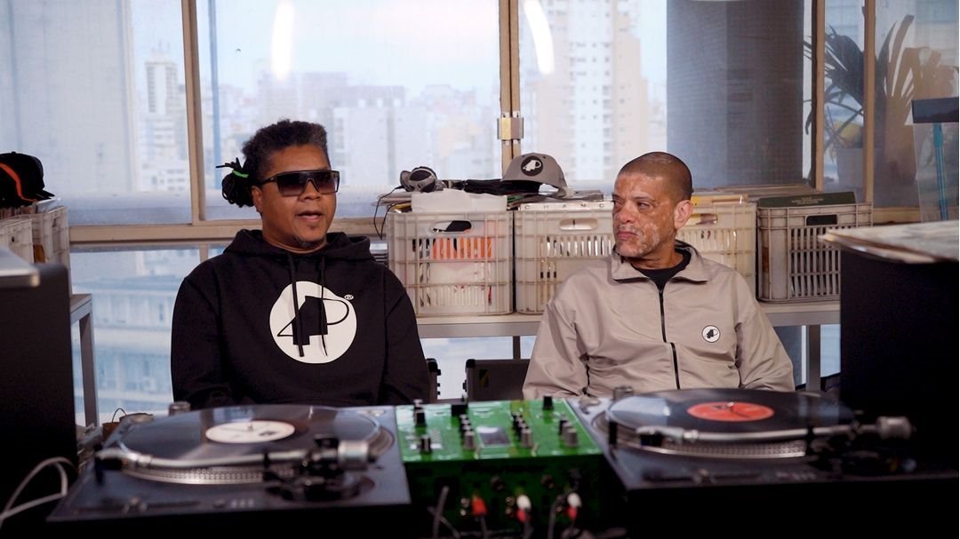 O rapper Xis e o DJ KL Jay, do Racionais MC's, em entrevista durante cena de um documentário. Os dois são negros e de meia idade. Xis usa um moletom preto e óculos escuros, enquanto KL Jay ysa um casaco bege de zipper. - Metrópoles