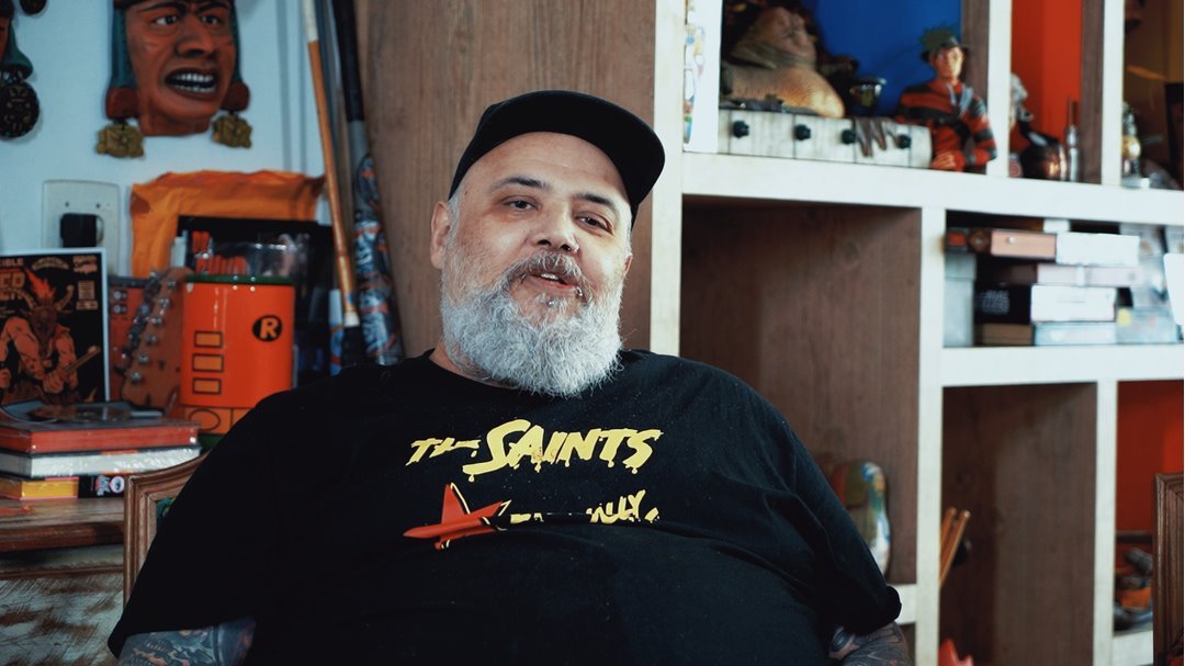 O vocalista da banda de punk Ratos de Porão, João Gordo, em entrevista ao documentário De Rua. Ele usa uma camiseta preta da banda de rock The Saints e um boné, também preto. João Gordo é um homem branco e de meia idade com barba branca longa. - Metrópoles