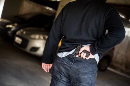 Imagem ilustrativa de homem guardando revólver na parte da trás da cintura enquanto anda num estacionamento - Metrópoles