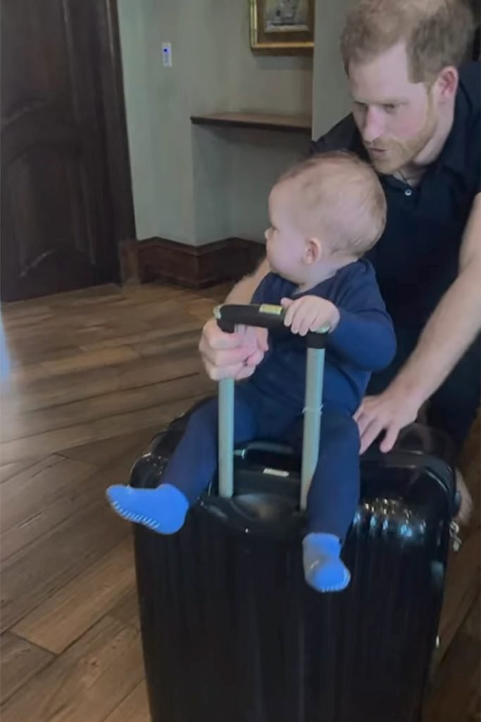 Foto colorida de bebê branco sentado em uma mala de viagem segurada por um adulto branco e ruivo - Metrópoles