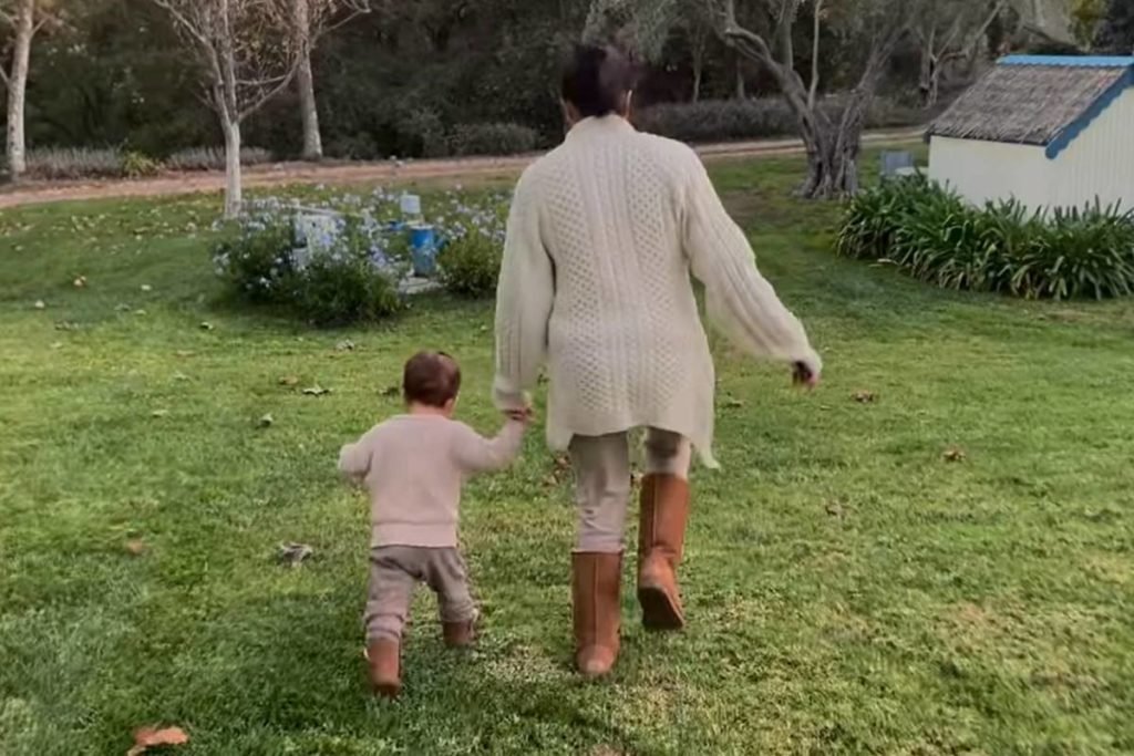 Foto colorida de menino e mulher de mãos dadas e de costas caminhando em um jardim - Metrópoles
