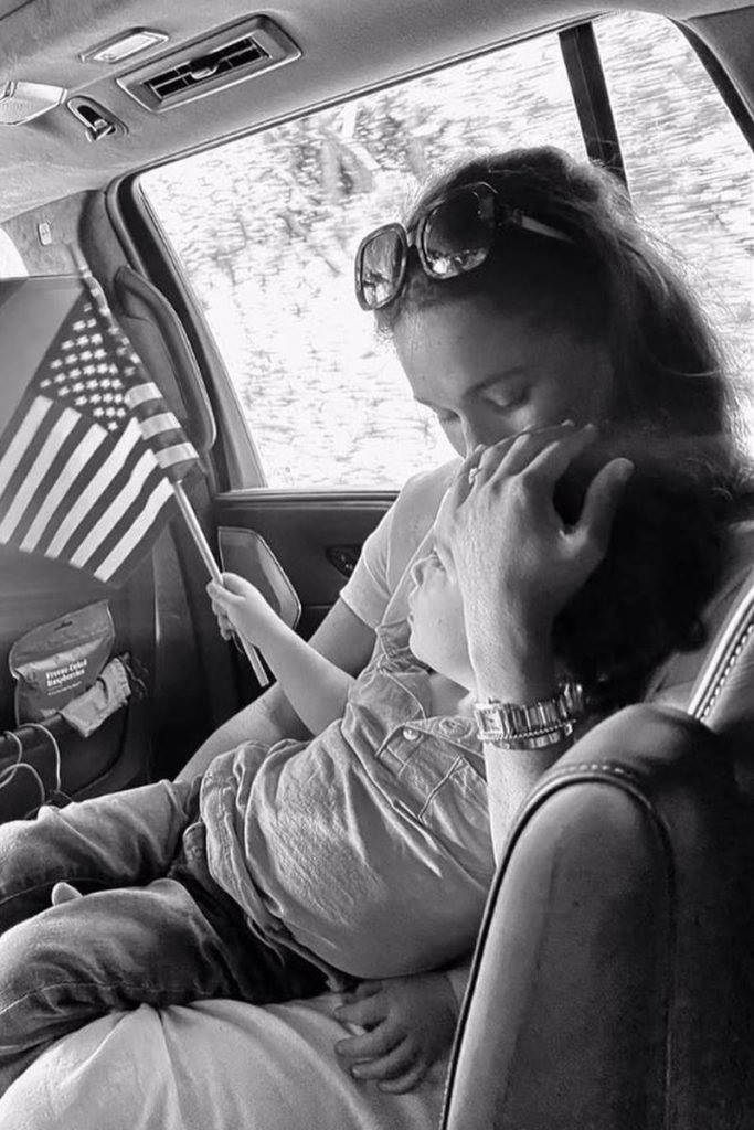 Foto em preto e branco de mulher dando beijo na testa de menino que segura bandeira dos Estados Unidos em uma das mãos - Metrópoles
