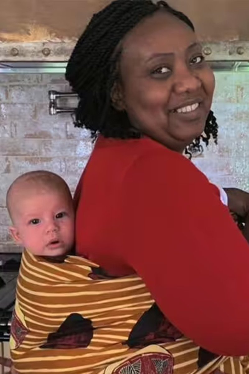 Foto colorida de bebê branco nas costas de uma mulher negra. Eles usam um suporte de pano - Metrópoles