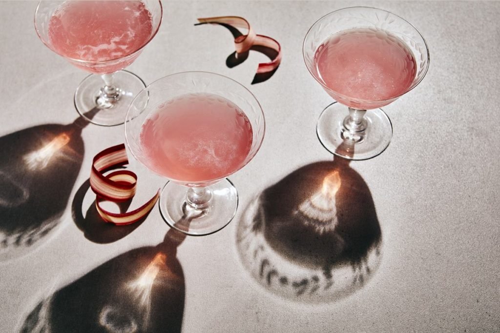Taça de vidro transparente com uma bebida rosa clara dentro. Na imagem é possível ver três taças com a bebida e pequenas balas, também cor de rosa. - Metrópoles