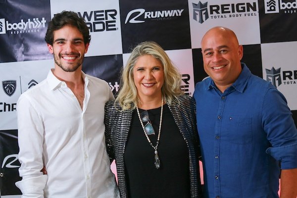 A man in a white dress shirt on the left, a blonde woman in a dark dress in the middle and a man with a blue dress shirt on the right in front of the launch poster for the new runway activity, Push and Play. 