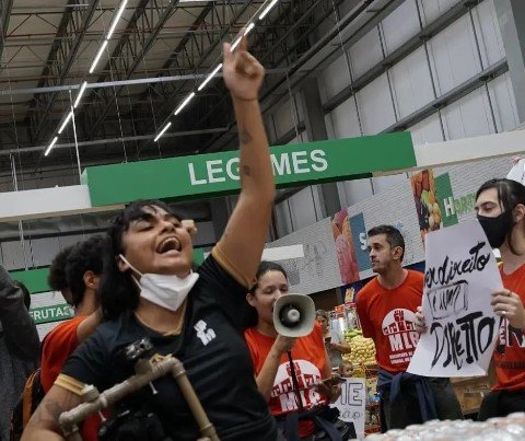 Mulher com máscara no queixo, camisa preta e mão levantada.  Atrás dela há pessoas de vermelho com cartazes e alto-falante.  Eles estão dentro de um supermercado
