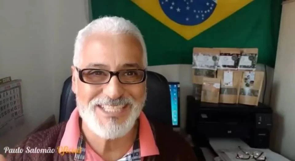 Homem com cabelo e barba brancos com óculos, sentado, sorrindo, e em frente a uma bandeira do Brasil
