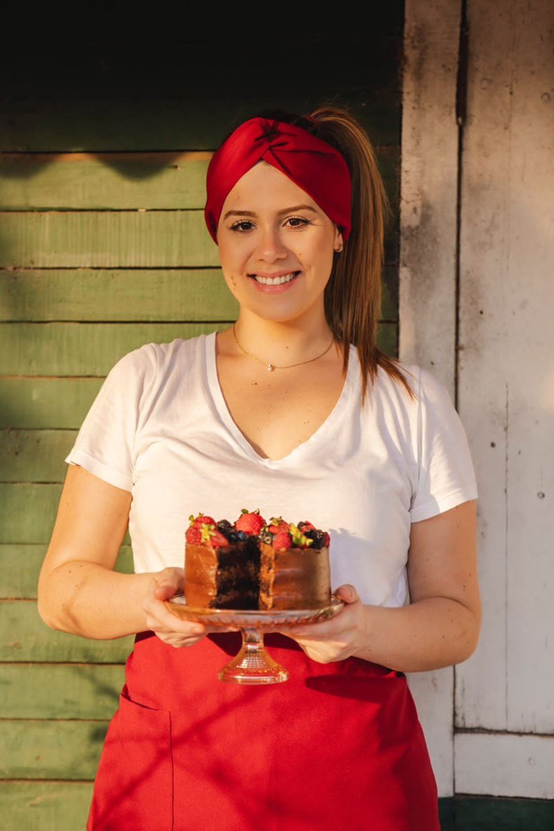 Foto colorida de mulher branca e loira segurando torta. Ela uma faixa no cabelo e avental na cor vermelha - Metrópoles