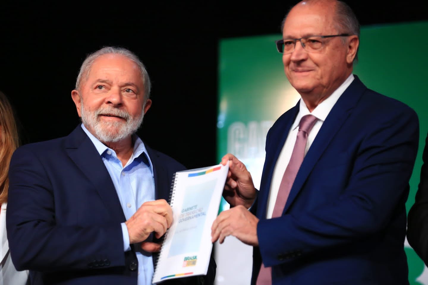 O vice-presidente eleito Alckmin e o presidente eleito Lula em coletiva de impresa para anúncio de novos ministros para o próximo governo. Eles seguram juntos o relatório da transição e sorriem - Metrópoles