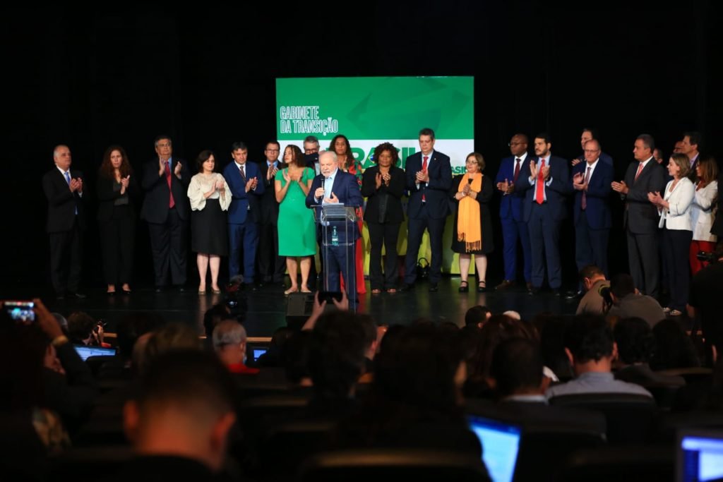 O presidente eleito Lula em coletiva de impresa para anúncio de novos ministros para o próximo governo. Ele fala de microfone na mão e com indicados aos ministérios atrás, o apludido - Metrópoles