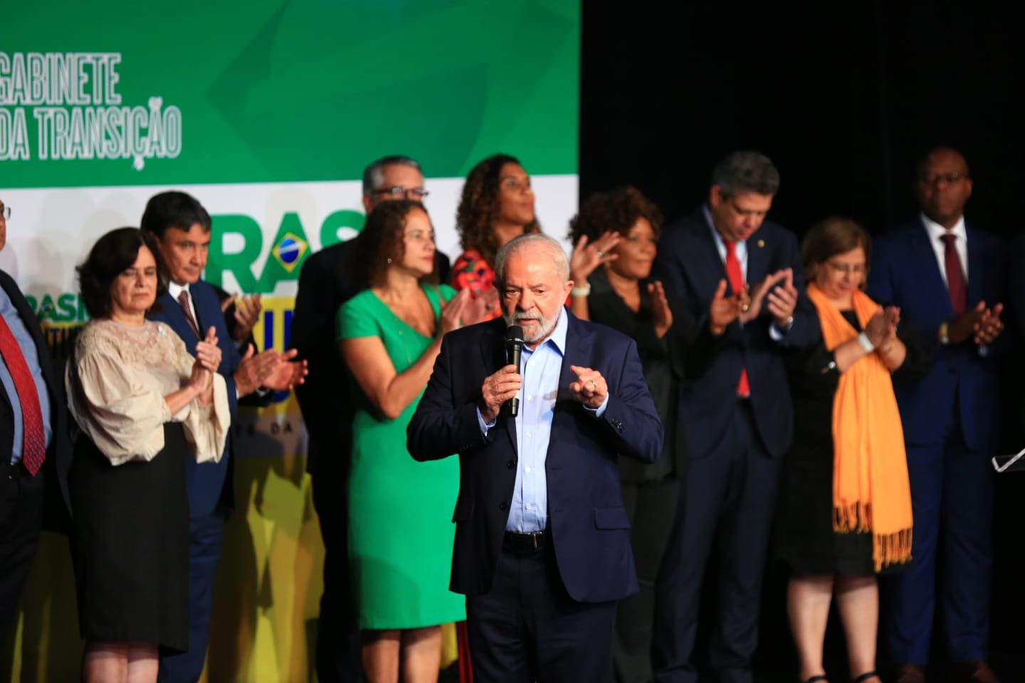 O presidente eleito Lula em coletiva de impresa para anúncio de novos ministros para o próximo governo. Ele fala de microfone na mão e com indicados aos ministérios atrás, o apludido - Metrópoles