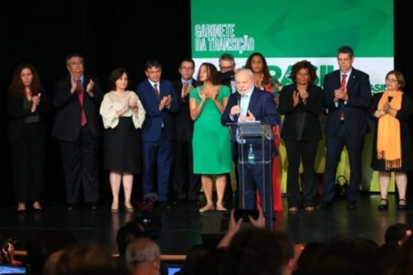 O presidente eleito Lula em coletiva de impresa para anúncio de novos ministros para o próximo governo. Ele fala de microfone na mão e com indicados aos ministérios atrás, o apludido - Metrópoles