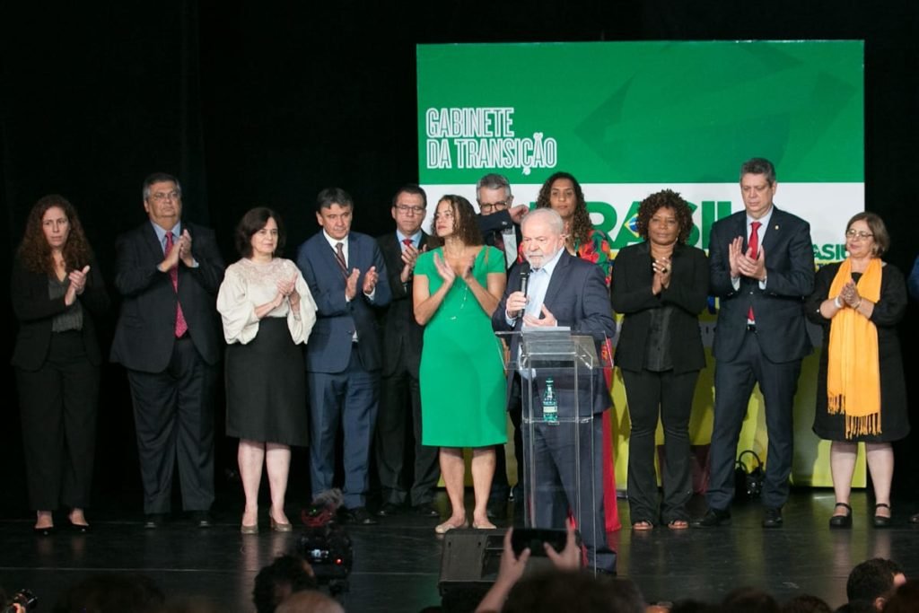 O presidente eleito Lula em coletiva de impresa para anúncio de novos ministros para o próximo governo. Ele fala de microfone na mão e com indicados aos ministérios atrás, o aplaudido - Metrópoles