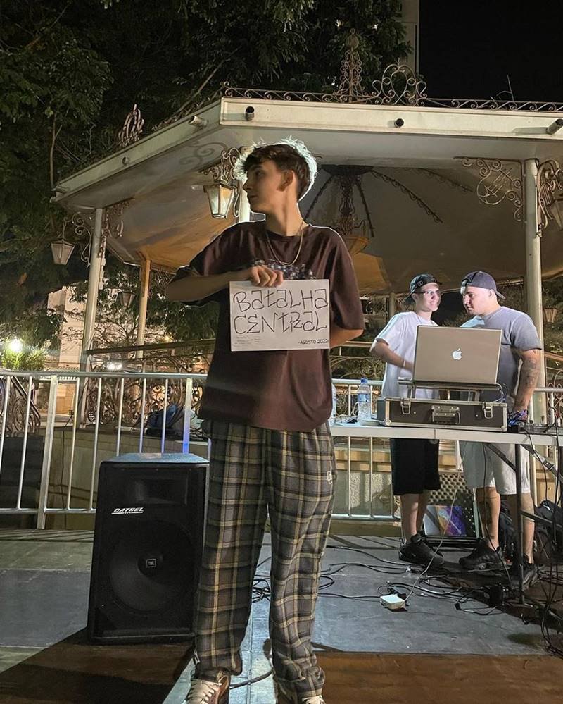 menino de calça zadrez e camiseta marrom segura placa escrito batalha central enquanto olha para o lado - metrópoles