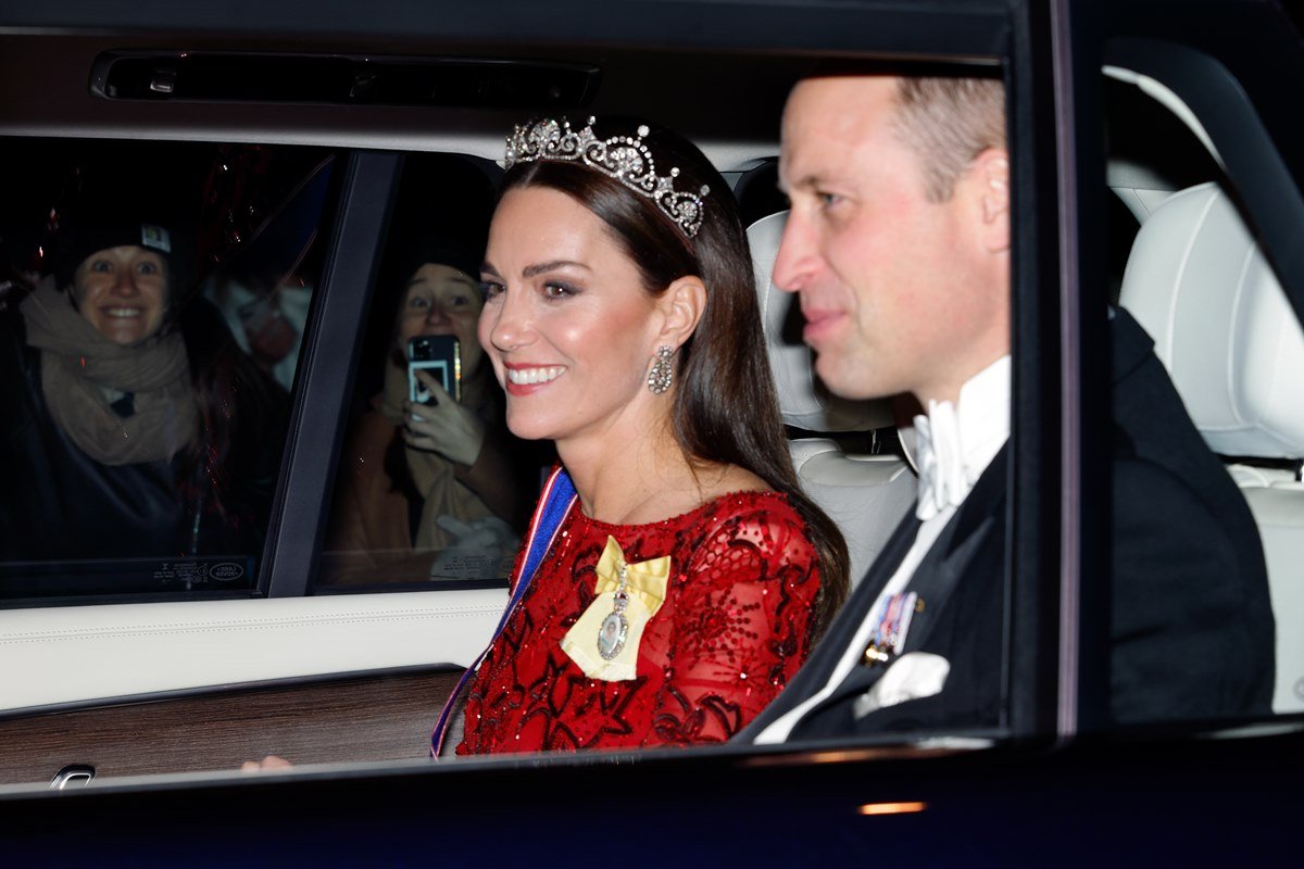 Foto colorida de mulher e homem brancos dentro de um carro. Ela usa uma tiara de pedras preciosas e uma roupa vermelha, enquanto ele está de terno - Metrópoles