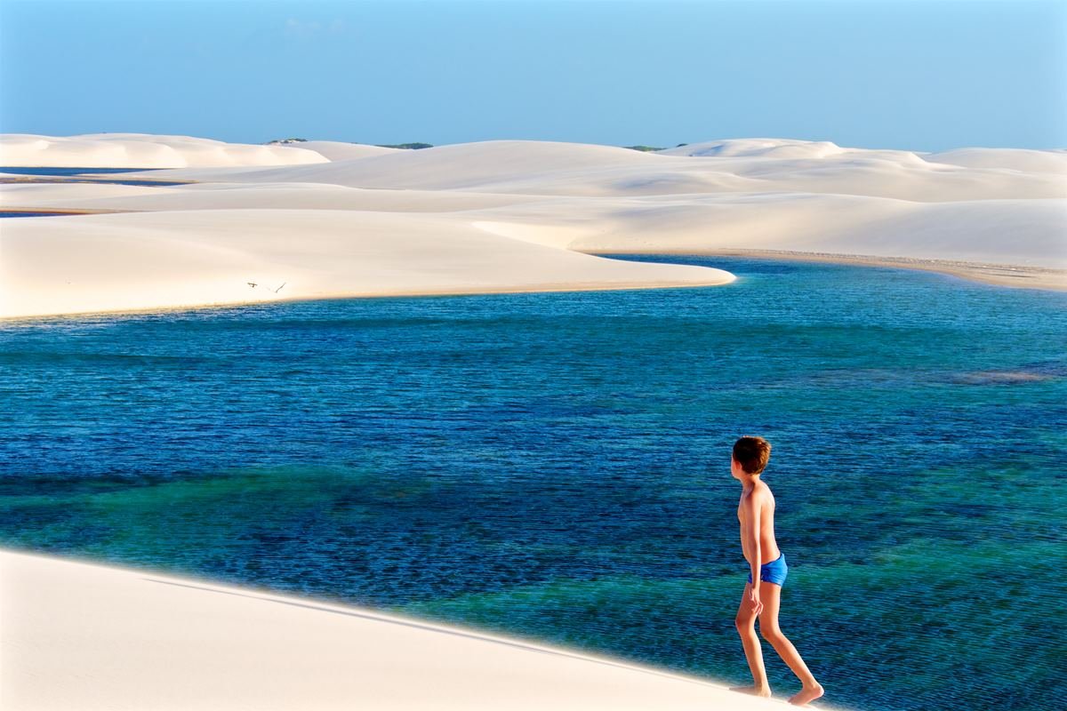 Foto colorida de menino subindo duna com água azul-turquesa - Metrópoles